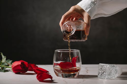 Crop unrecognizable person in white shirt pouring whiskey from crystal jar into transparent glass with petals placed on table near rose flower