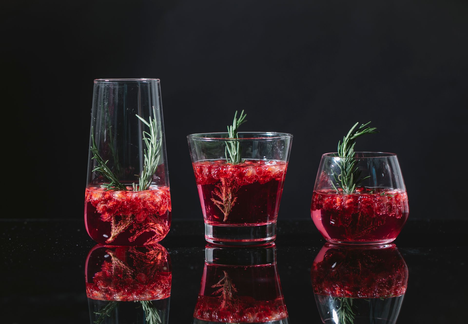 Transparent clean glasses filled with similar red cocktail with rosemary and berries on black background