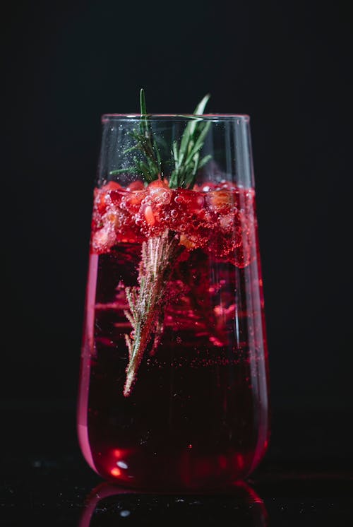 Crystal glass of alcohol cocktail with pomegranate berries and rosemary twig against black background