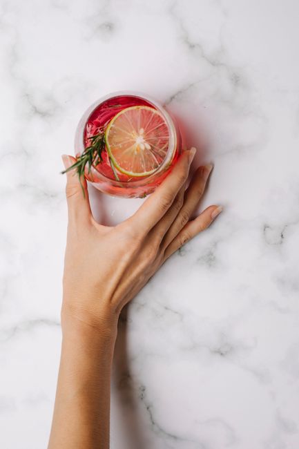 From above of crop anonymous woman taking glass of tasty cocktail with slices of lemon and sprig of rosemary against light marble surface