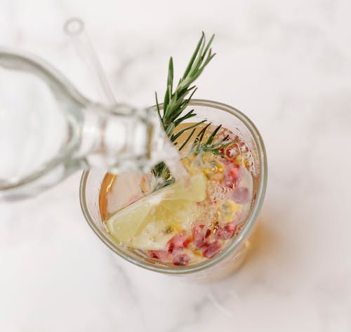 From above of glass bottle pouring water into glass with sprig of rosemary and slice of lemon placed on marble surface