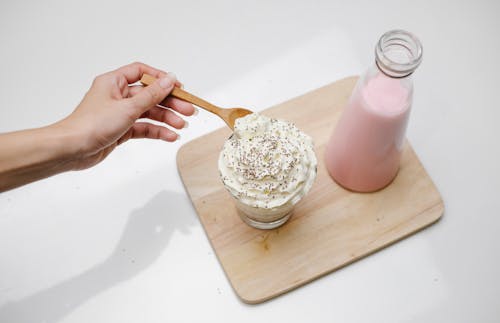 High angle of crop unrecognizable woman checking by wooden teaspoon froth of whipped cream on dessert served near bottle with pink milkshake on wooden board