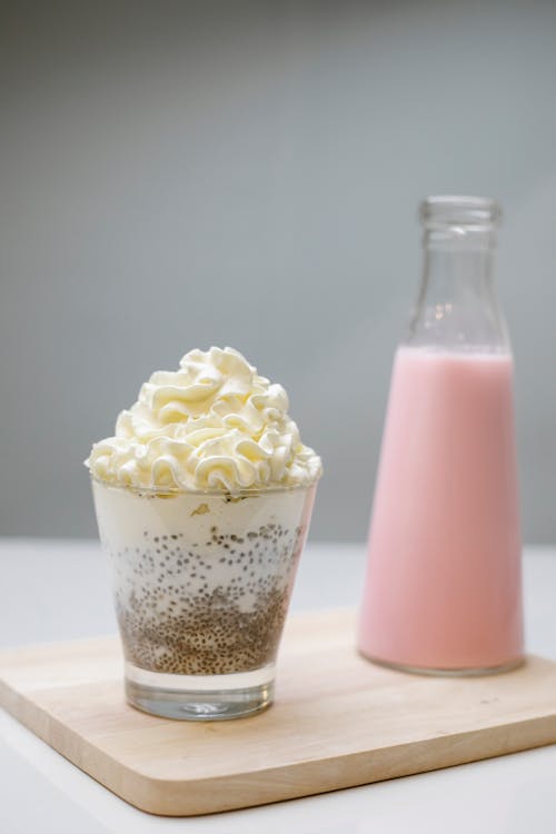 Side view of portion bottle with tasty pink milkshake and glass with milk pudding with chia seeds decorated whipped cream served wooden board on gray background