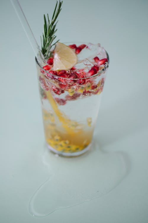 From above glass of tasty fresh cold drink with pomegranate seeds and passion fruit pure decorated rosemary and lemon and added transparent straw on white table in puddle