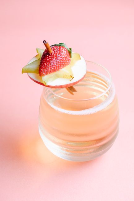 High angle of wide transparent glass with fruit cocktail decorated with slices of strawberry starfruit and apple on cinnamon stick placed on pink surface