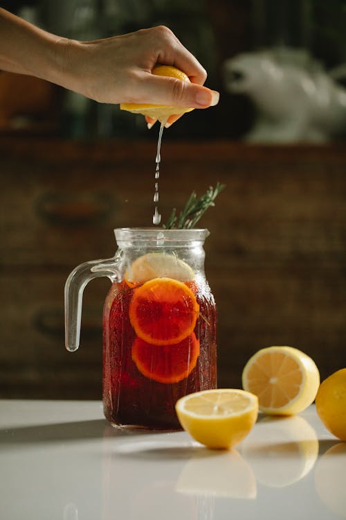 Crop anonymous female squeezing half of ripe lemon into glass jar with homemade refreshing drink with lemon slices and rosemary