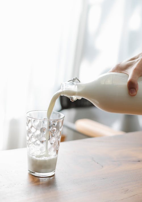 Crop faceless person pouring fresh cold milk from bottle into transparent clean glass placed on wooden table
