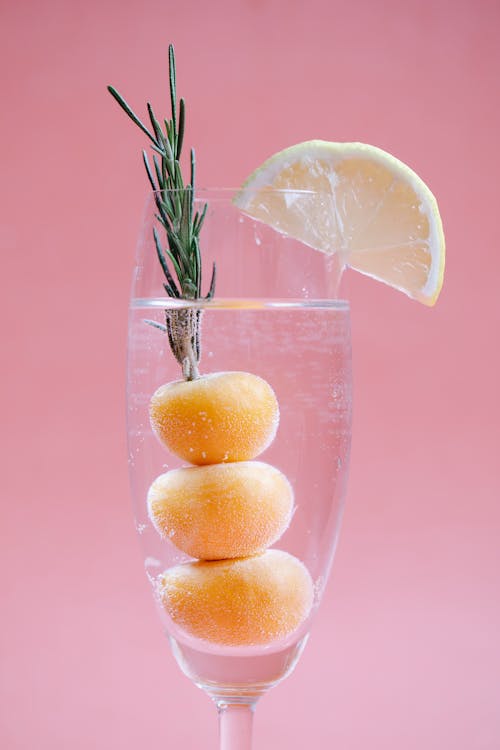 Transparent clean glass of refreshing cocktail with apricots and rosemary decorated with slice of lemon on pink background