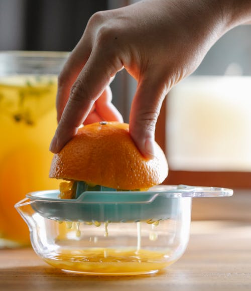 Crop anonymous person squeezing unpeeled half of ripe orange while making fresh juice in kitchen