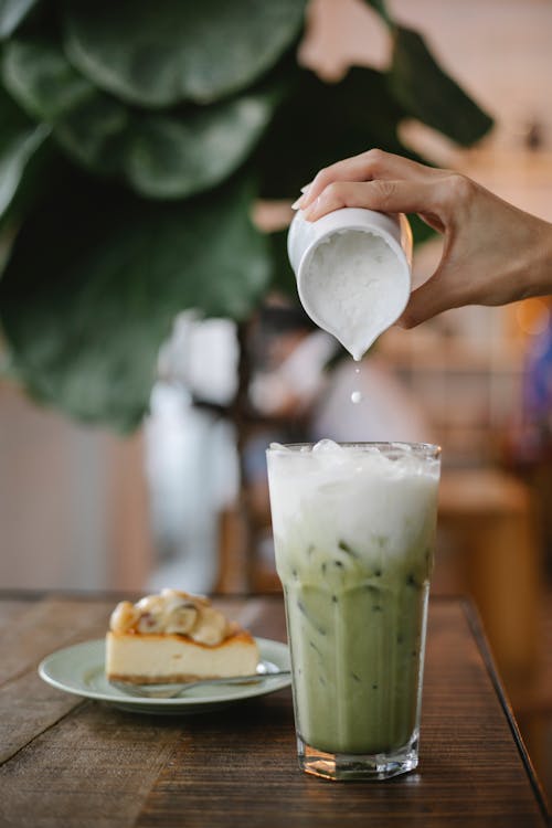Free Crop anonymous female pouring whipped milk into glass with matcha frappe placed on table near plate with dessert in cafe Stock Photo