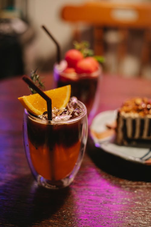 Gelas Minum Bening Dengan Cairan Coklat Dan Irisan Buah Di Atas Piring Keramik Putih