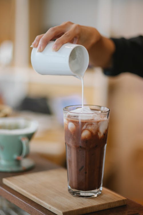 Crop anonymous female barista adding whipped milk in transparent glass with ice coffee placed on wooden board