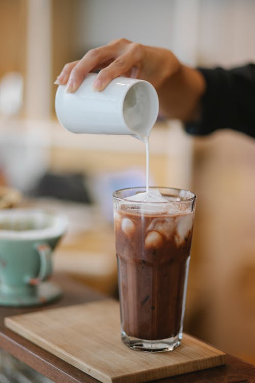 Crop anonymous female barista adding whipped milk in transparent glass with ice coffee placed on wooden board