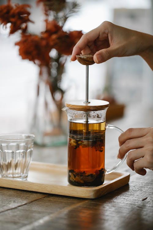 Free Crop anonymous female brewing hot aromatic herbal tea in glass French press placed on wooden tray on table in cafe Stock Photo