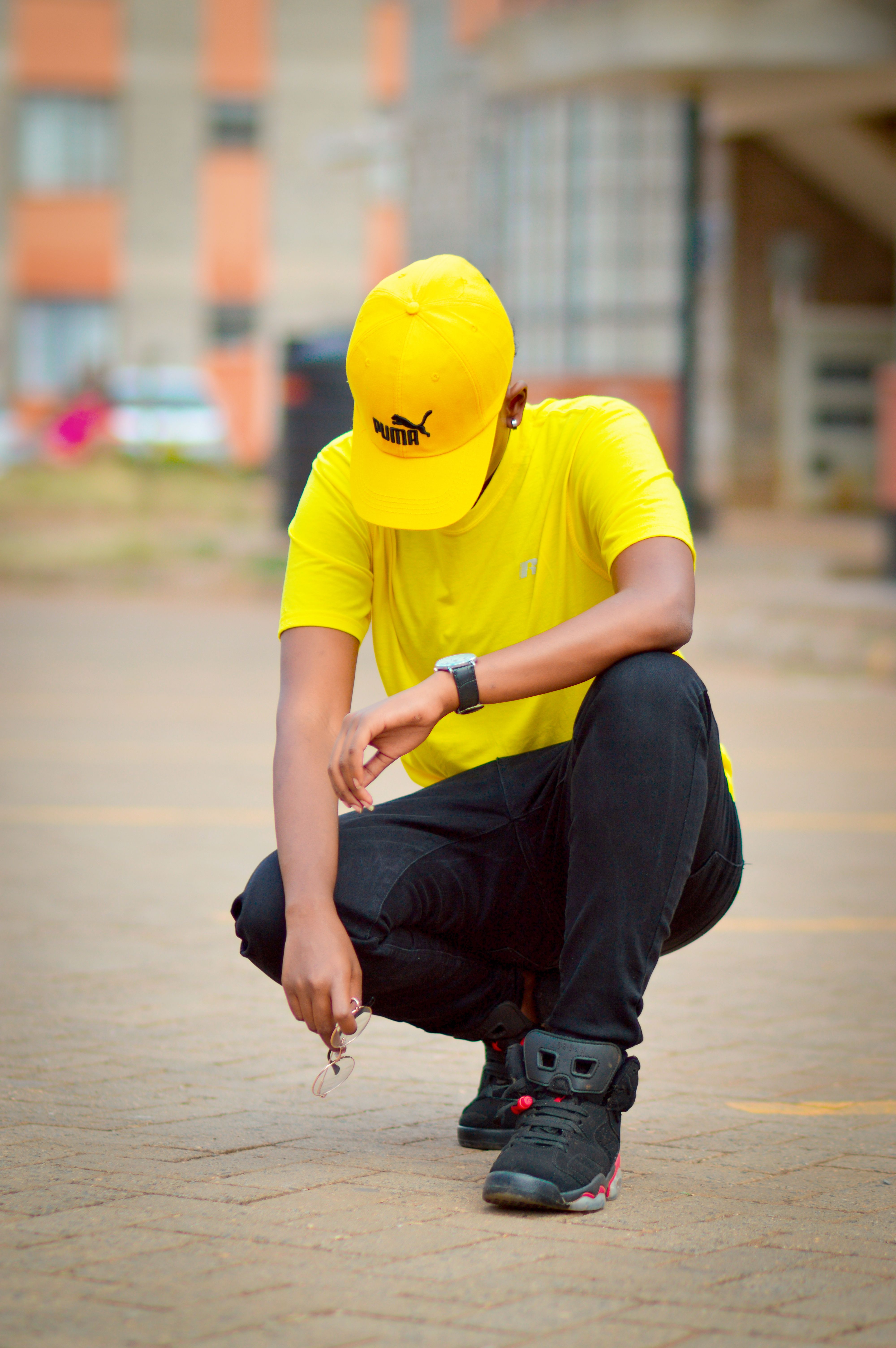 Stylish individual in yellow cap and shirt, black pants, kneeling outdoors capturing modern street style.