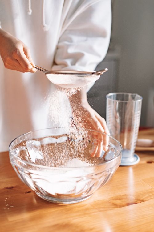 Free A person is sieving flour into a glass bowl for baking preparation on a wooden table. Stock Photo