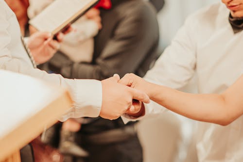 Unrecognizable priest with bible holding hands of anonymous bride and groom while taking vows  during wedding ceremony with guests on blurred background