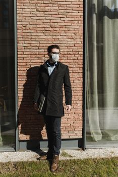 白天，一个戴着面具和眼镜的高加索人拿着笔记本电脑站在砖墙前