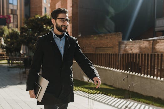 Businessman walking outdoors with a smile, holding a laptop, exuding confidence.