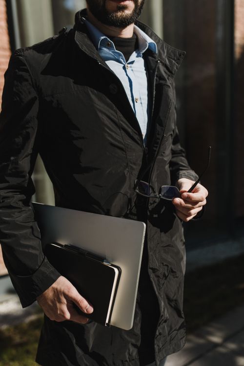 Free Businessman outdoors holding laptop and notebook, dressed in black jacket. Stock Photo