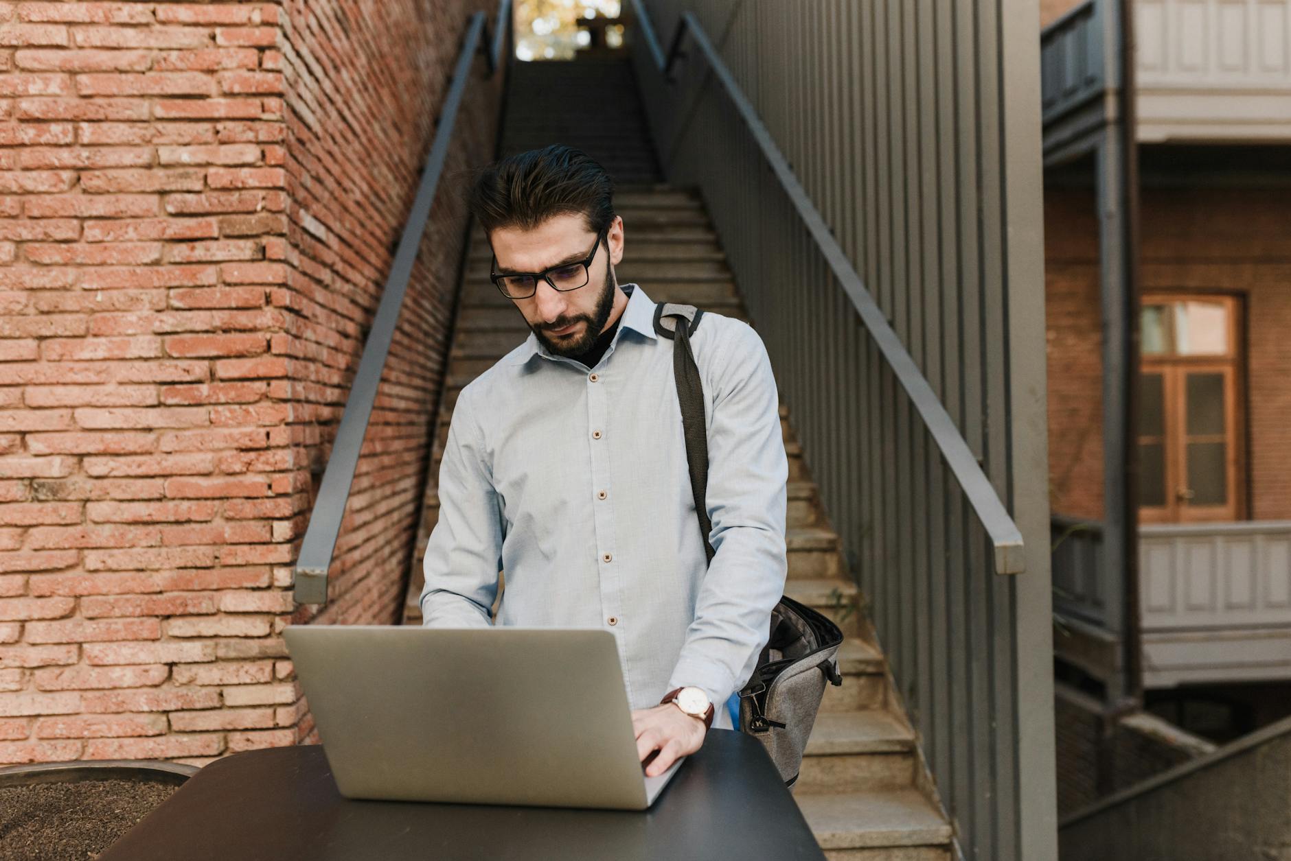 A man with glasses works on a laptop outdoors near brick stairs, depicting remote work lifestyle.