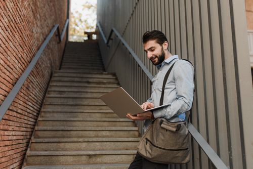 Free Businessman working remotely on laptop, leaning against a wall outdoors. Stock Photo