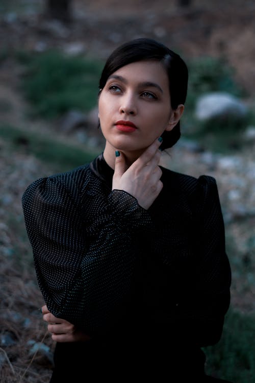 Young attractive female wearing stylish black blouse touching neck tenderly and looking away dreamily while standing in green nature