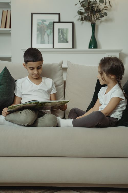 Free Two children sitting on a sofa, reading a book together, creating a cozy and familial atmosphere. Stock Photo