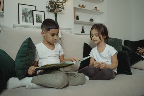 Free A young boy and girl reading a book together on a sofa with plush pillows in a warm, family setting. Stock Photo