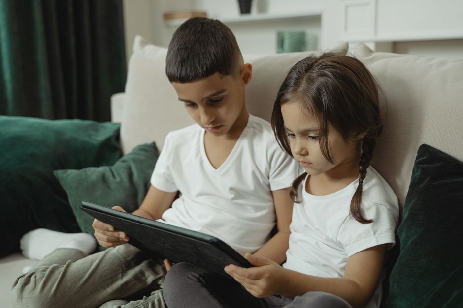 Two children focused on a tablet while sitting together on a sofa indoors.