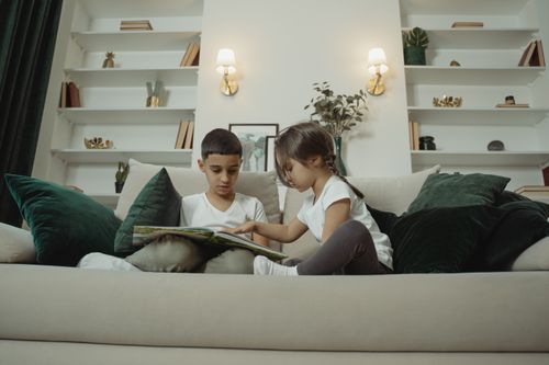 Free Two children sitting on a sofa reading a book together in a modern living room. Stock Photo