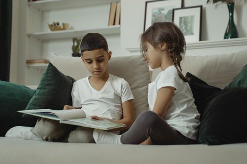 Free A young girl and boy reading a book on a comfortable sofa, surrounded by pillows. Stock Photo