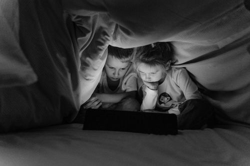 Free Two children watching a tablet under a blanket in a cozy bedroom setting, in grayscale for an intimate mood. Stock Photo