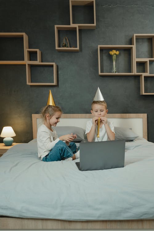 Little Brother and Sister Wearing Birthday Hats and Sitting on a Bed with a Laptop 