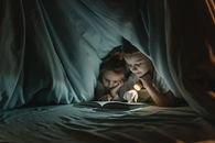 Two children read a book under a blanket using a flashlight, showcasing a cozy night in.