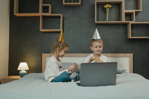 Free Little Brother and Sister Wearing Birthday Hats and Sitting on a Bed with a Laptop  Stock Photo