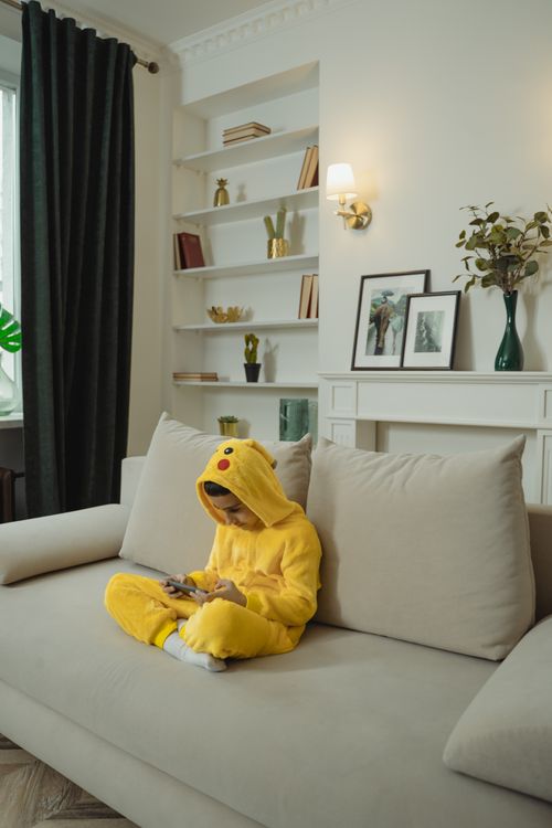 Free Young boy in a Pikachu costume using a smartphone while sitting on a sofa in a cozy living room. Stock Photo