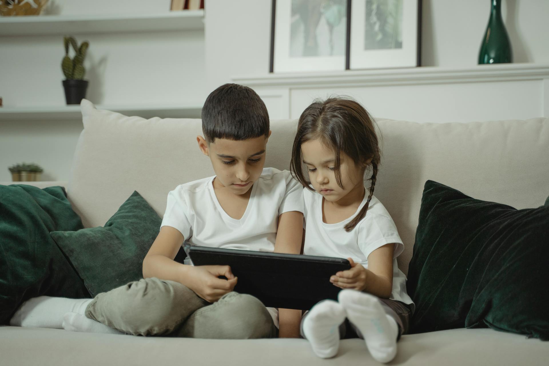 Two children sitting on a sofa sharing a digital tablet, creating a cozy indoor atmosphere.