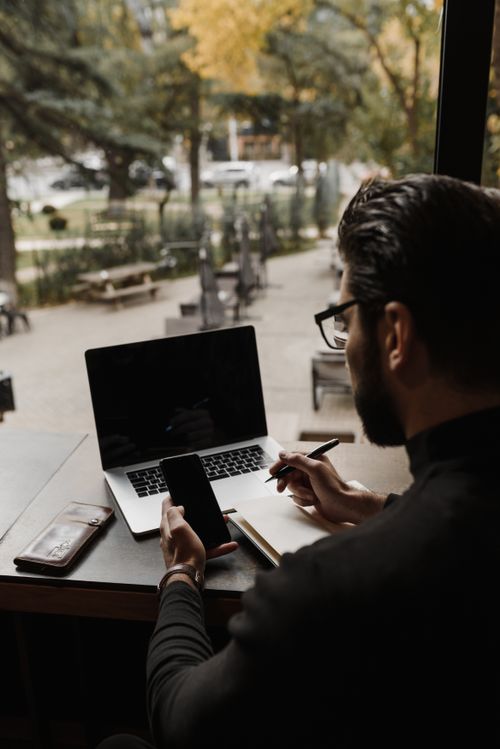 Free A man working remotely with a laptop and smartphone in a cozy indoor setting with a view of a park. Stock Photo