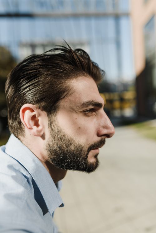 Free Side profile of a bearded man in a light blue shirt outdoors, thoughtful expression. Stock Photo