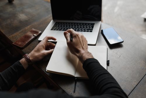 Free Overhead view of hands writing in a notebook beside a laptop and smartphone on a wooden desk. Stock Photo