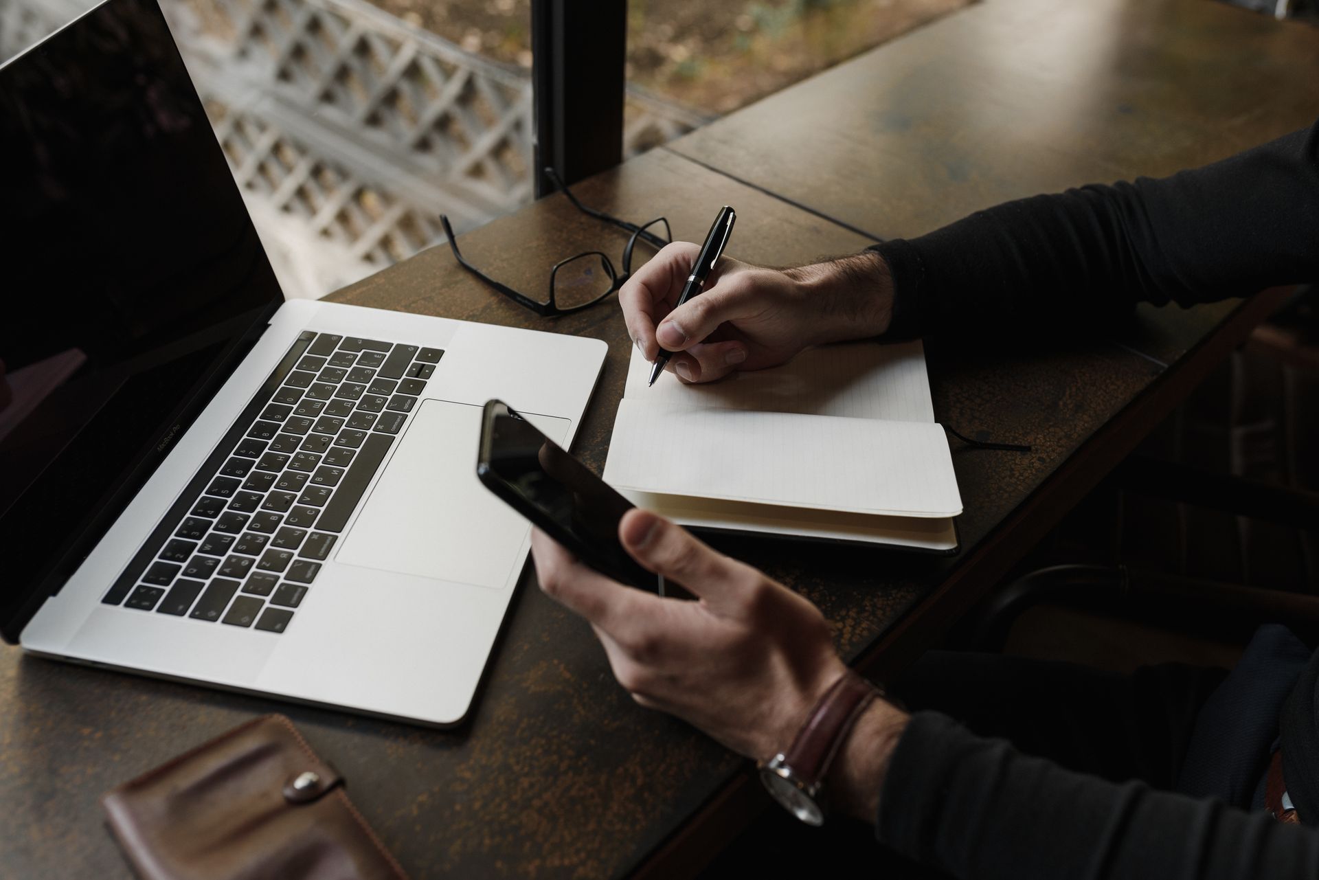 A business professional multitasking with a laptop, smartphone, and notebook in a modern workspace.
