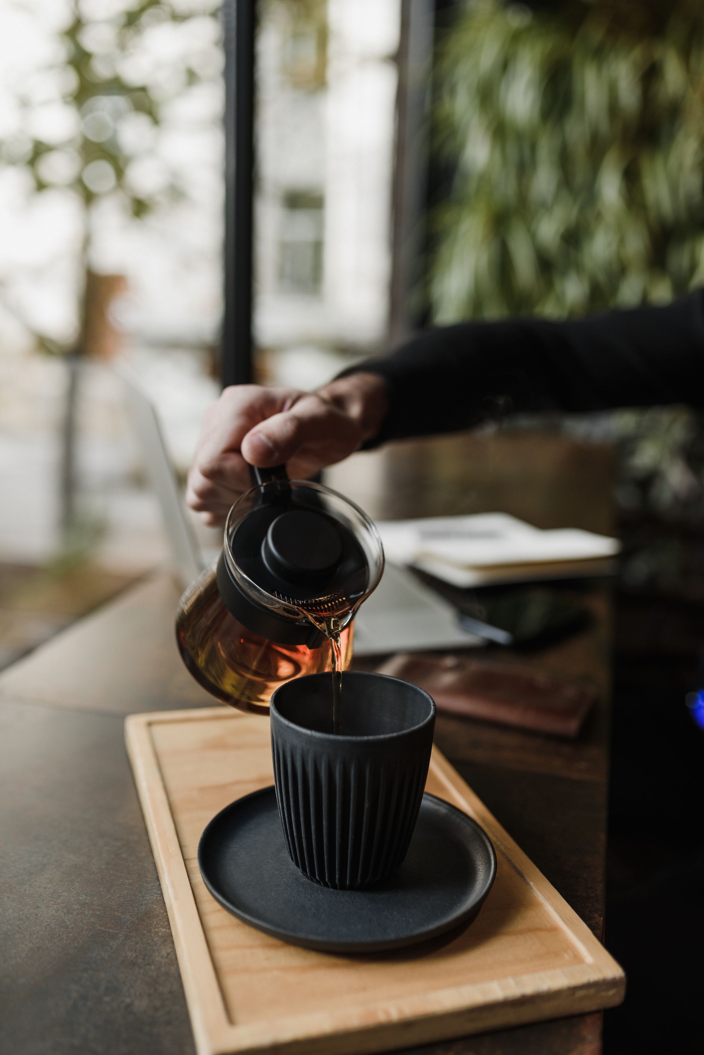 A person pours hot coffee into a stylish black mug on a wooden tray, creating a cozy vibe.