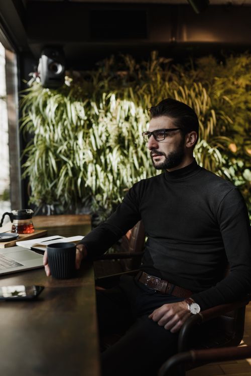 Free Bearded man with eyeglasses holding a cup, seated at a table in a cozy indoor cafe. Stock Photo