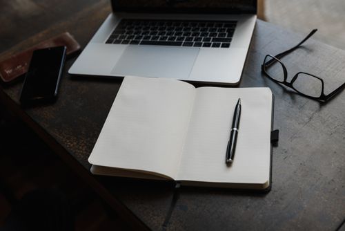 Free A workspace featuring a laptop, open notebook, pen, and eyeglasses on a wooden desk. Stock Photo