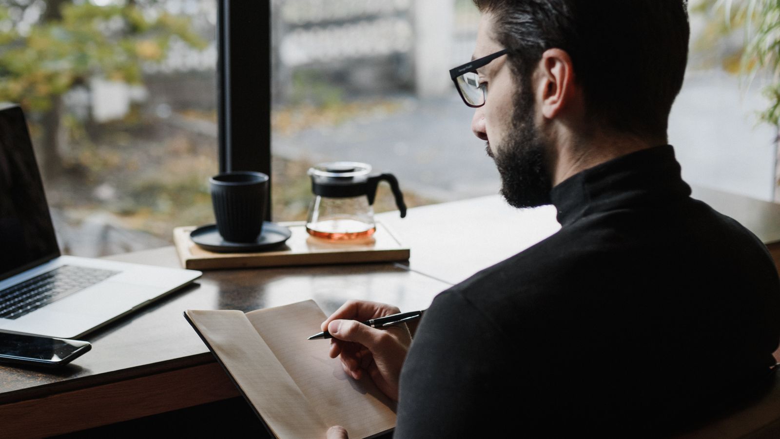 An adult writing in a notebook at a table beside a window