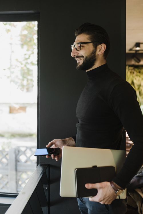 Free Confident businessman holding a laptop and smartphone, standing by a window indoors. Stock Photo