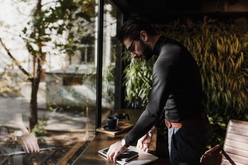 Free Businessman in a café standing by a window with sunlight, working on a laptop. Stock Photo