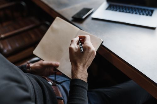 Free Close-up of a person writing in a notebook at a wooden desk with a laptop and phone. Stock Photo