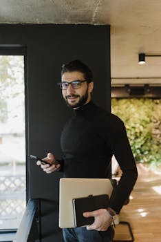 A man with a beard and eyeglasses holding a laptop and smartphone indoors.